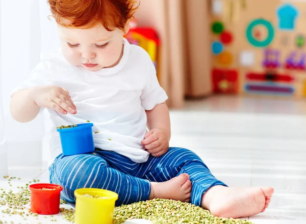 Cute redhead baby boy developing his fine motility skills by playing with green peas at home — Stock Photo, Image