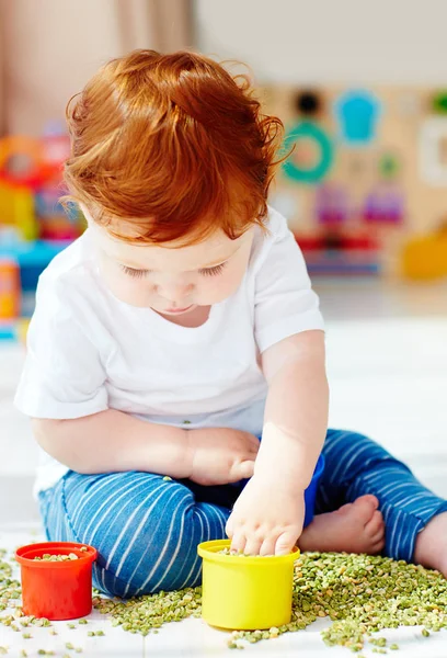 Carino rossa bambino ragazzo sviluppare la sua multa motilità abilità giocando con i piselli verdi a casa — Foto Stock