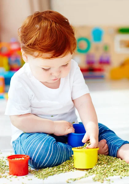 Bonito ruiva bebê menino desenvolvendo suas habilidades de motilidade fina, jogando com ervilhas verdes em casa — Fotografia de Stock