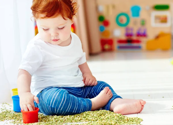 Lindo pelirrojo bebé niño desarrollando sus habilidades de motilidad fina jugando con guisantes verdes en casa —  Fotos de Stock