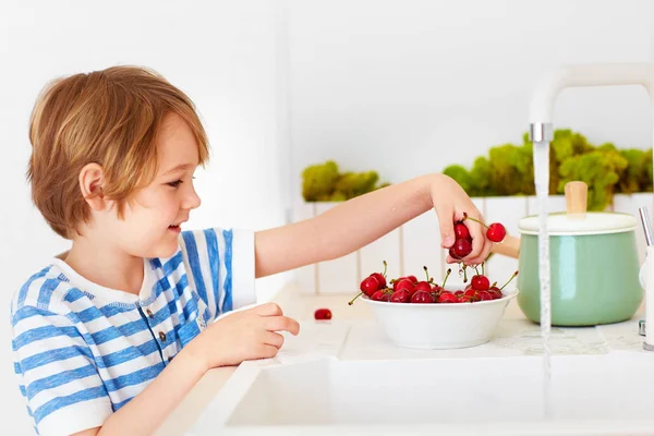 Schattige jonge jongen wassen van de armful van zoete kersen onder water van de kraan in de keuken — Stockfoto