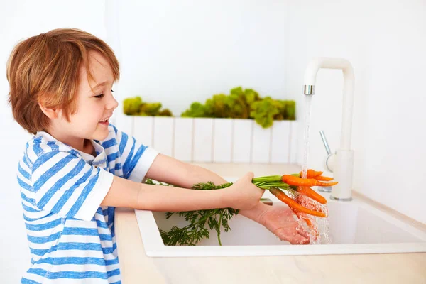 Menino bonito lavar as cenouras sob água da torneira na cozinha — Fotografia de Stock