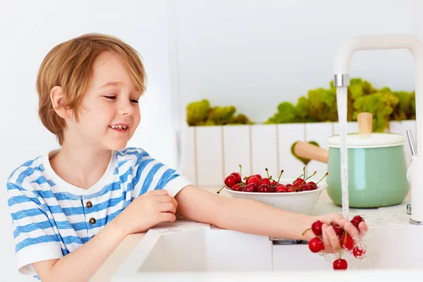 Schattige jonge jongen wassen van de armful van zoete kersen onder water van de kraan in de keuken — Stockfoto