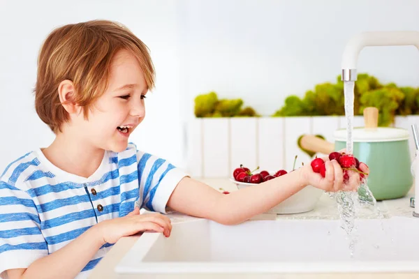 Anak muda yang bahagia mencuci setumpuk buah ceri manis di bawah air keran di dapur — Stok Foto