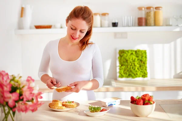 Glad ung kvinna förbereder läckra snacks vid köksbordet i morgonljuset — Stockfoto