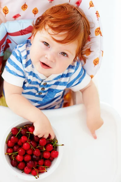 Bebê ruiva bonito degustação de cerejas doces enquanto sentado em cadeira alta na cozinha — Fotografia de Stock