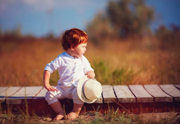 Αστεία κοκκινομάλλα toddler αγοράκι κάθεται στο ξύλινο μονοπάτι στο πεδίο του καλοκαιριού — Φωτογραφία Αρχείου