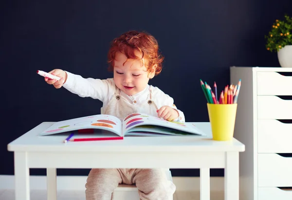 Feliz pelirroja bebé dibujo en el desarrollo de libro en el escritorio — Foto de Stock