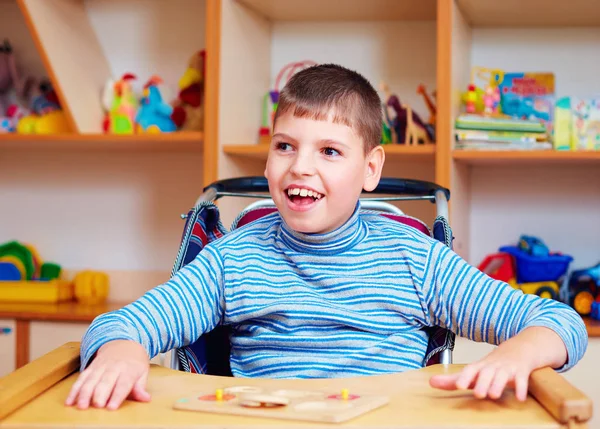 Menino alegre com deficiência no centro de reabilitação para crianças com necessidades especiais, resolvendo quebra-cabeça lógico — Fotografia de Stock