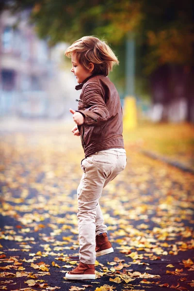 Lindo niño, chico en chaqueta de cuero que se divierte en la calle de otoño, saltando y corriendo sobre la alfombra de hojas caídas —  Fotos de Stock