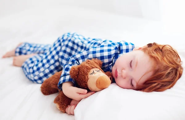 Adorable pelirroja bebé pequeño durmiendo con peluche juguete en pijama de franela —  Fotos de Stock