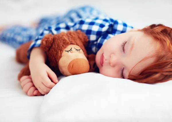 Entzückendes rothaariges Kleinkind im Flanell-Pyjama, das mit Plüschwärmer schläft — Stockfoto