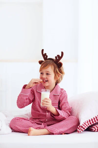 Menino feliz bonito, criança em pijama de flanela tradicional sentado na cama com copo de leite fresco e biscoitos, feriados de Natal — Fotografia de Stock