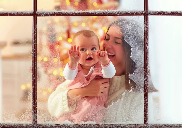 Linda niña bebé en la mano madre mirando a través de la ventana, vacaciones de invierno familiares —  Fotos de Stock