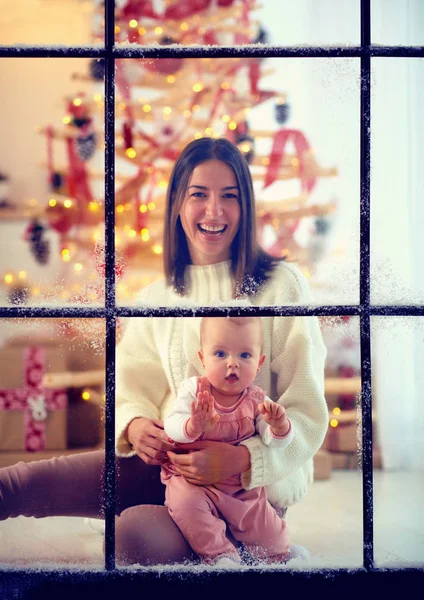 Lycklig mamma och baby flicka njuter snörik vinter genom fönstret, julhelgen — Stockfoto