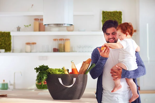 Feliz padre ofreciendo a bebé hijo una fruta fresca de la cesta de la compra, cocina casera — Foto de Stock