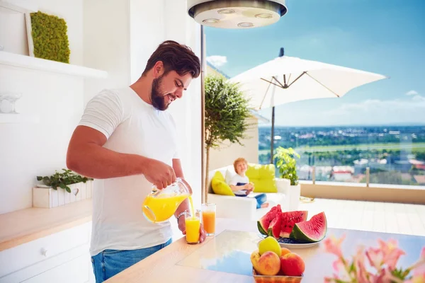 Hombre adulto joven, padre vertiendo jugo fresco mientras está de pie en la cocina de espacio abierto en un día soleado de verano — Foto de Stock