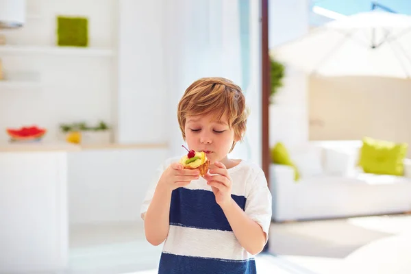Cute kid, young boy eating tasty cupcake with whipped cream and fruits at home — Stock Photo, Image