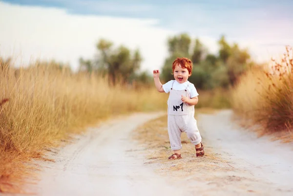 Αξιολάτρευτο κοκκινομάλλα αγοράκι μικρό παιδί σε ολόσωμη φόρμα που τρέχει κατά μήκος του δρόμου αγροτικές καλοκαίρι ηλιοκαμένος πεδίο — Φωτογραφία Αρχείου