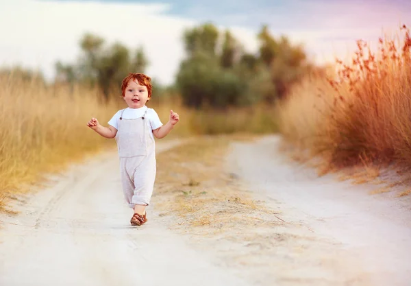 Прелестный рыжий малыш в комбинезоне бегает по сельской дороге на загорелом поле — стоковое фото
