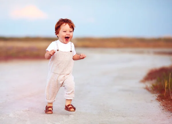 Adorable redhead toddler baby boy in jumpsuit running through the summer road and field — Stock Photo, Image