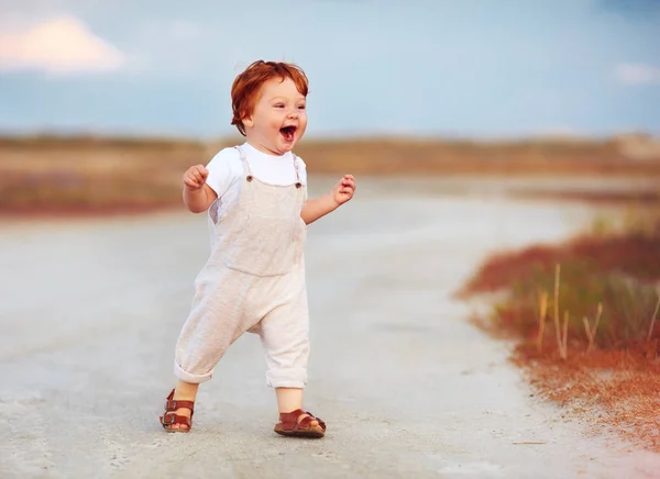 Adorable pelirroja bebé niño en mono corriendo a través de la carretera de verano y el campo —  Fotos de Stock