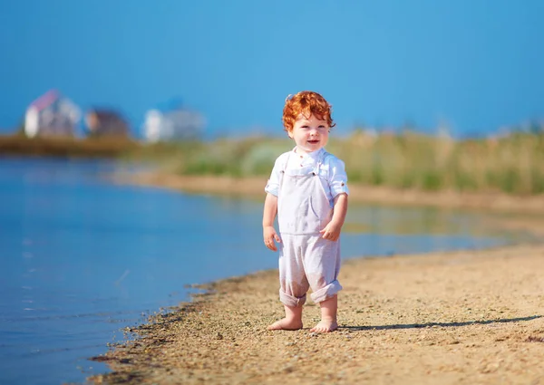 Roztomilá zrzka batole chlapeček chůzi na pobřeží jezera v letní ráno — Stock fotografie