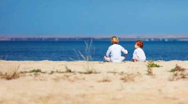 Dos niños pequeños, hermanos sentados en la playa de arena en la mañana cerca del lago —  Fotos de Stock