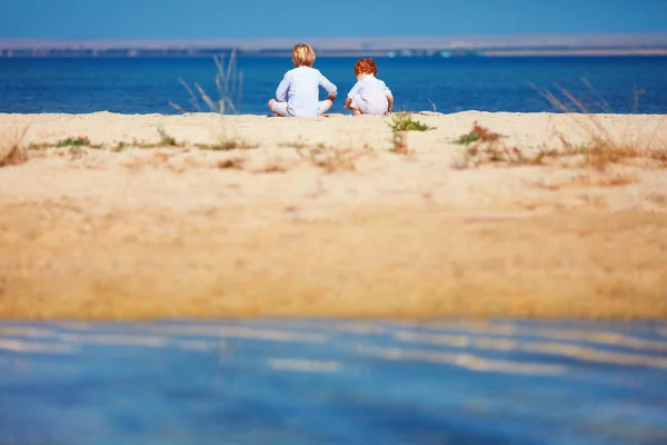 Δύο νεαρά παιδιά, αδέλφια κάθονται σε αμμώδη παραλία το πρωί κοντά στη λίμνη — Φωτογραφία Αρχείου