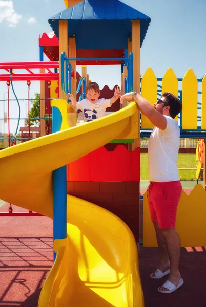 Happy excited toddler baby boy sliding in colorful playground at summer day with the help of father — Stock Photo, Image