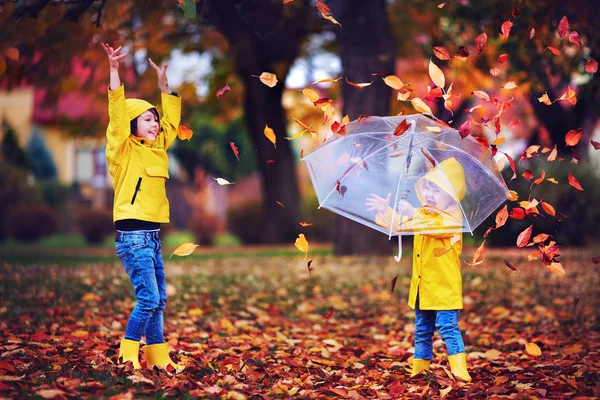 Glücklich aufgeregte Kinder, die Spaß haben, Blätter im Herbstpark werfen, Regenstiefel und Mäntel tragen — Stockfoto