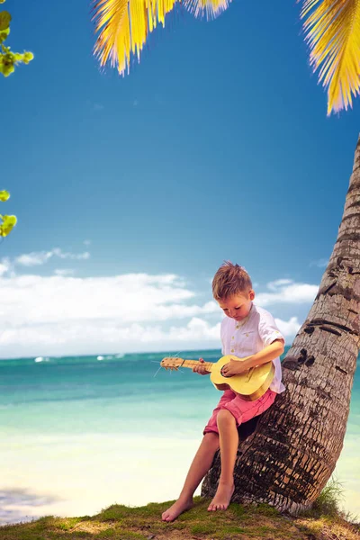 Cute kid, young boy playing guitar, ukulele on summer tropical beach under the palm tree — Stock Photo, Image