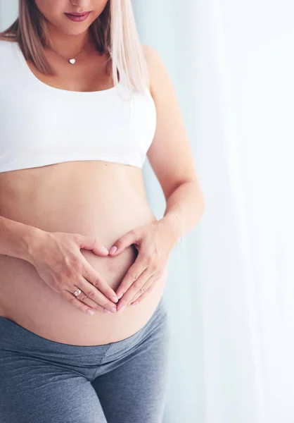 Happy pregnant woman placing hands on belly in heart symbol — Stock Photo, Image