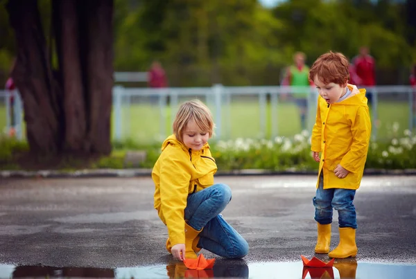 Ragazzi carini, fratelli che si divertono insieme, lanciando le barche di carta nelle pozzanghere primaverili, indossando impermeabili e stivali di gomma — Foto Stock