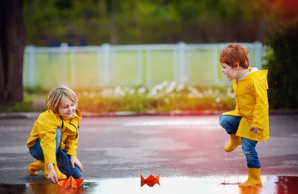 Leuke jongens, broers hebben plezier samen, de lancering van de papieren boten in het voorjaar plassen, het dragen van regenjassen en rubberen laarzen — Stockfoto