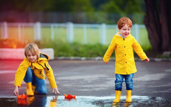 Ragazzi carini, fratelli che si divertono insieme, lanciando le barche di carta nelle pozzanghere primaverili, indossando impermeabili e stivali di gomma — Foto Stock