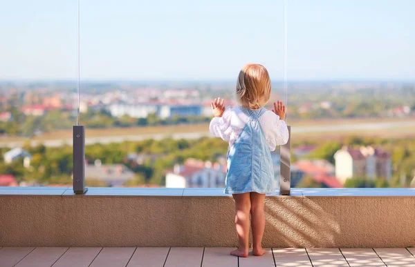 Mignon tout-petit bébé fille regardant le paysage de la ville à travers le verre balustade au patio sur le toit — Photo