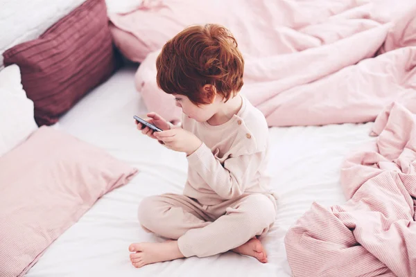 Lindo pelirrojo bebé niño en pijama jugando videojuegos en el teléfono en el dormitorio por la mañana —  Fotos de Stock
