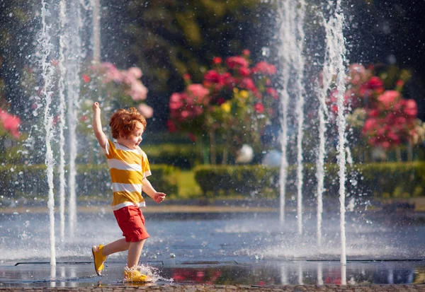 Opgewonden roodharige kleine jongen heeft plezier tussen waterstralen, in fontein. Zomer in de stad — Stockfoto