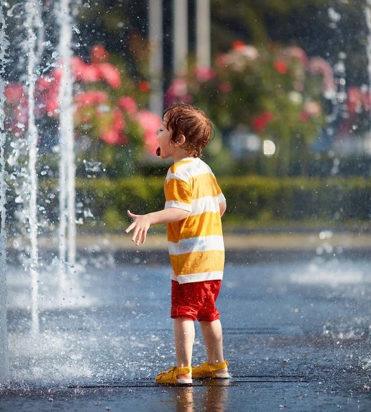 Schattig roodharige baby jongen hebben plezier tussen fontein water jets, het vangen van het water druppels op zijn tong — Stockfoto