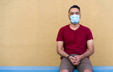 Karantina sırasında yüz maskesi takan yakışıklı, çekici bir Asyalı adam, kendini tecrit ve tecrit, sarı-kahverengi arka planda, kırmızı tişörtte oturuyor.
