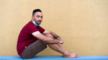 Karantina sırasında yüz maskesi takan yakışıklı, çekici bir Asyalı adam, kendini tecrit ve tecrit, sarı-kahverengi arka planda, kırmızı tişörtte oturuyor.