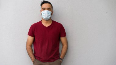 Karantina sırasında yüz maskesi takan yakışıklı, çekici bir Asyalı adam, tecrit ve tecrit, gri arka planda duruyor, kırmızı tişörtlü, sıkılmış, mutsuz.