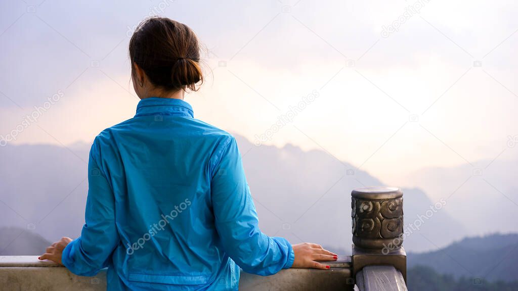 Young brunette woman from the back is standing on the peak of the mountain at the national park and watching a sunrise, beautiful travel concept, 