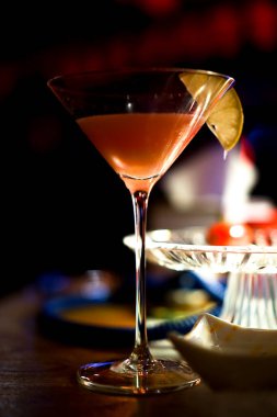 Uzun martini bardağı, pembe kokteyl ve bir dilim limon karanlık gece kulübünde, seçici bir odak noktası.