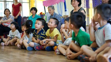  Bir grup küçük Asyalı çocuk yerde oturuyor, bir anaokulundaki öğretmenlere bakıyor ve onları dinliyorlar.