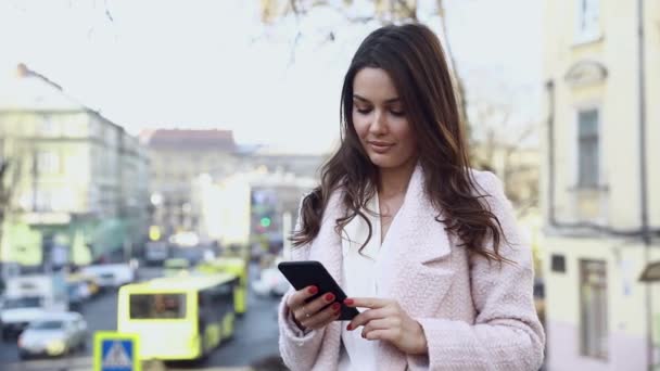 通りに立っている間 彼女のスマートフォンにメッセージを入力する魅力的な女性 楽しい時間をアウトドアを楽しんでいます カメラの前でスマートフォンとかなりブルネット 美しい女の子 — ストック動画