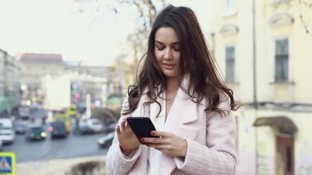 一个迷人的女人站在街上用她的智能手机打字 玩的开心 在户外享受时光 漂亮的黑发 智能手机在相机前 漂亮的女孩 — 图库视频影像