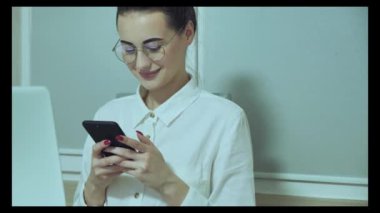 Adorable European girl with laptop computer on table while texting on smart phone online chat with friend. Elegant young office woman in suit sitting  in the office working waiting for meeting customers.