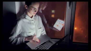 Young self employed woman working in the office sitting on the windowsill by the large window. business woman working on a laptop late at night in the office on a background of night city lights. Her raises his head and laughs into the camera.